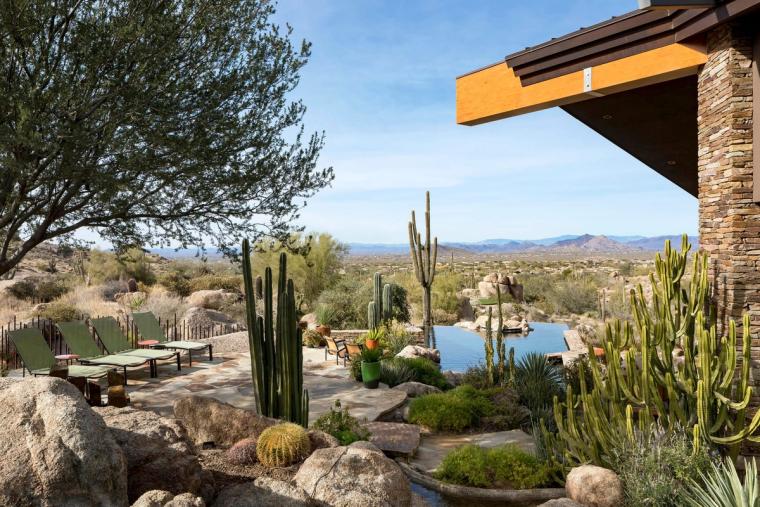 View from a shaded stone-and-timber patio out over cactus planting and a pond to open desert and distant hills