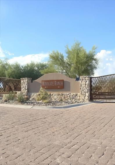 Stone and stucco entry monument lettered WINDGATE RANCH beside a paver drive and metal gate, desert planting around it