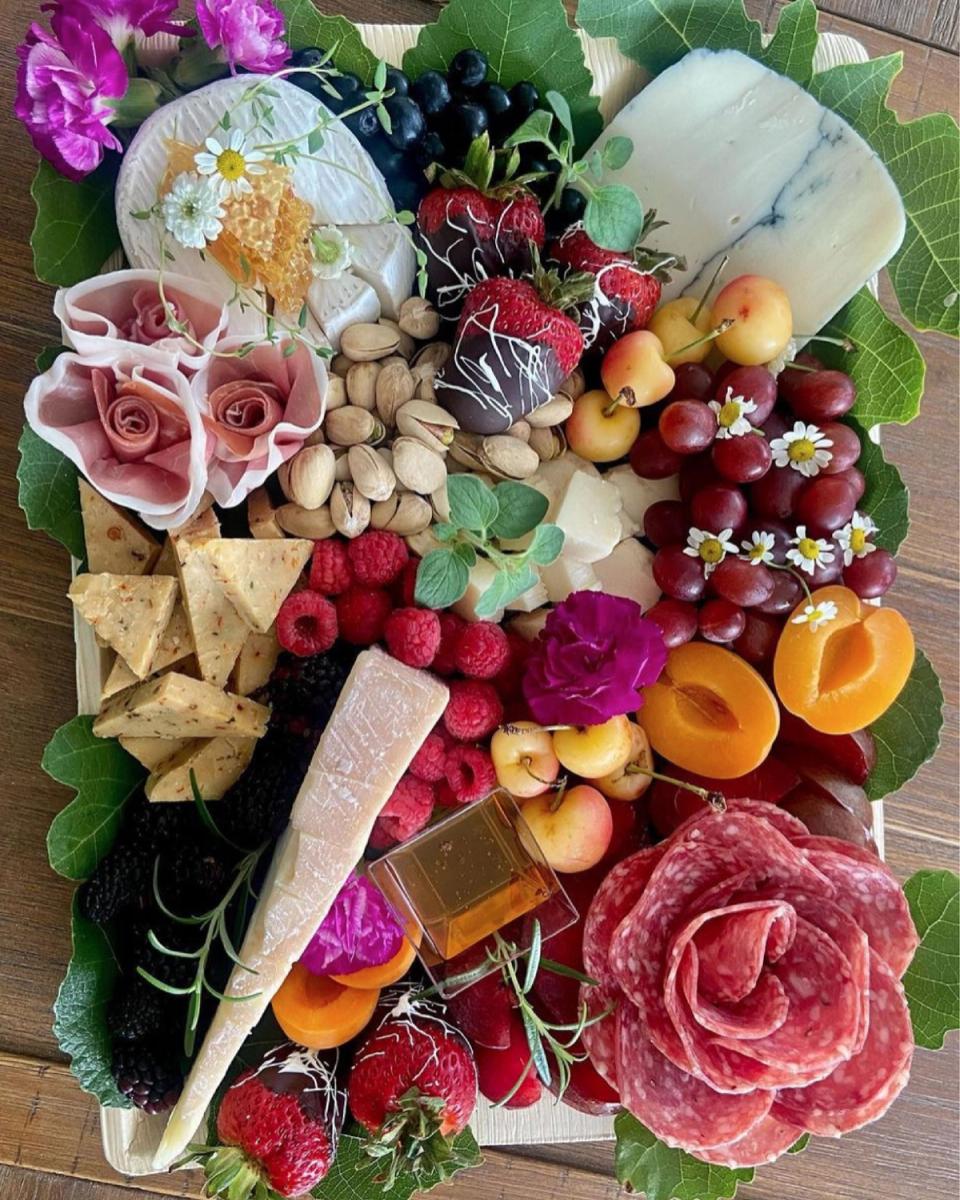 Overhead view of a charcuterie board with brie, blue cheese, berries, stone fruit, pistachios, a rolled prosciutto rose, and edible flowers on grape leaves