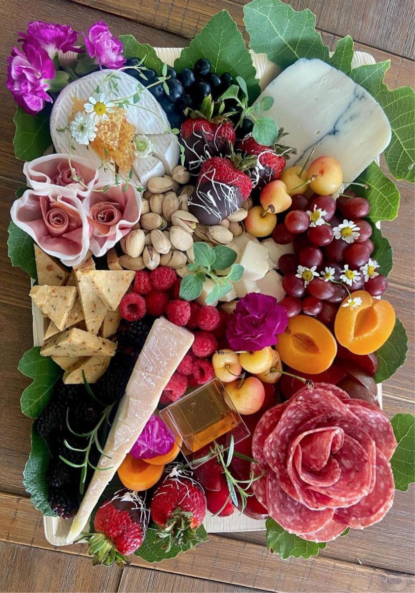 Overhead view of a charcuterie board with brie, blue cheese, berries, stone fruit, pistachios, a rolled prosciutto rose, and edible flowers on grape leaves