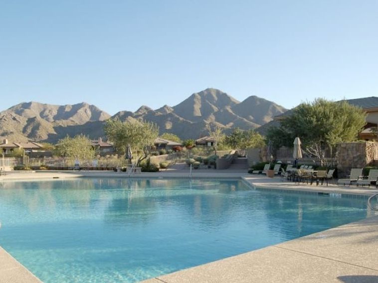 Community swimming pool with handrails and rows of loungers, ramadas along the far edge and a jagged mountain range beyond