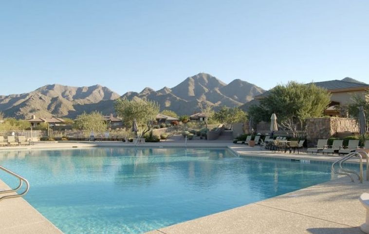 Community swimming pool with handrails and rows of loungers, ramadas along the far edge and a jagged mountain range beyond