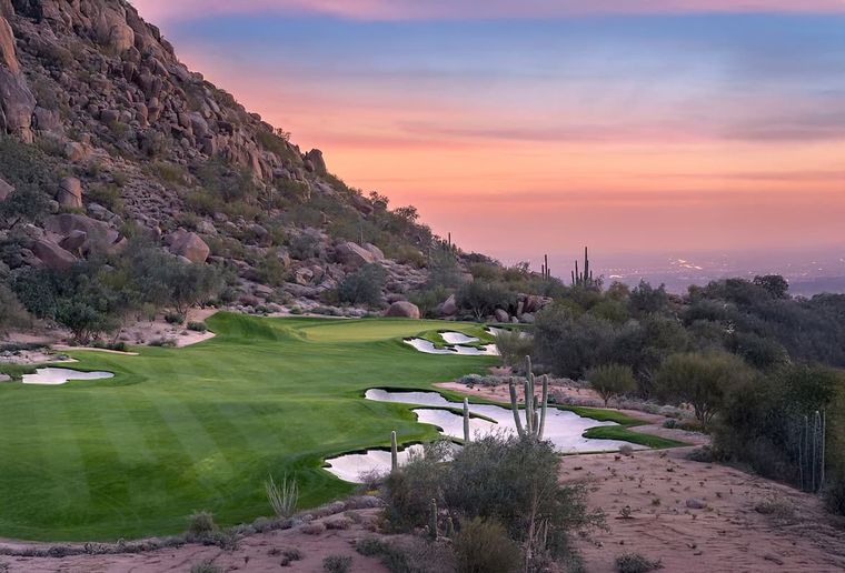 Desert golf holes and bunkers at dusk below a boulder ridge, distant city lights