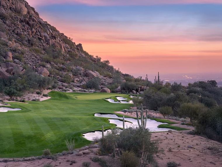 Desert golf holes and bunkers at dusk below a boulder ridge, distant city lights