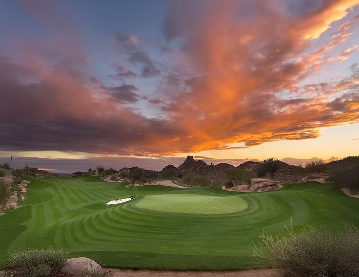 Golf green and flagstick under a vivid orange sunset over granite desert boulders
