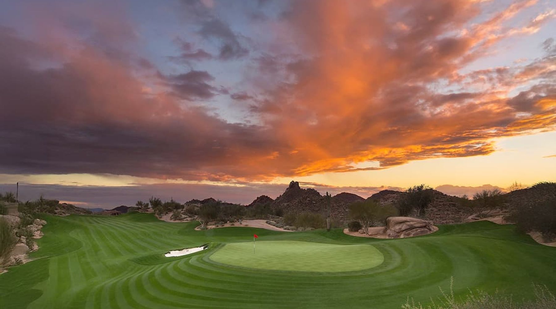 Golf green and flagstick under a vivid orange sunset over granite desert boulders