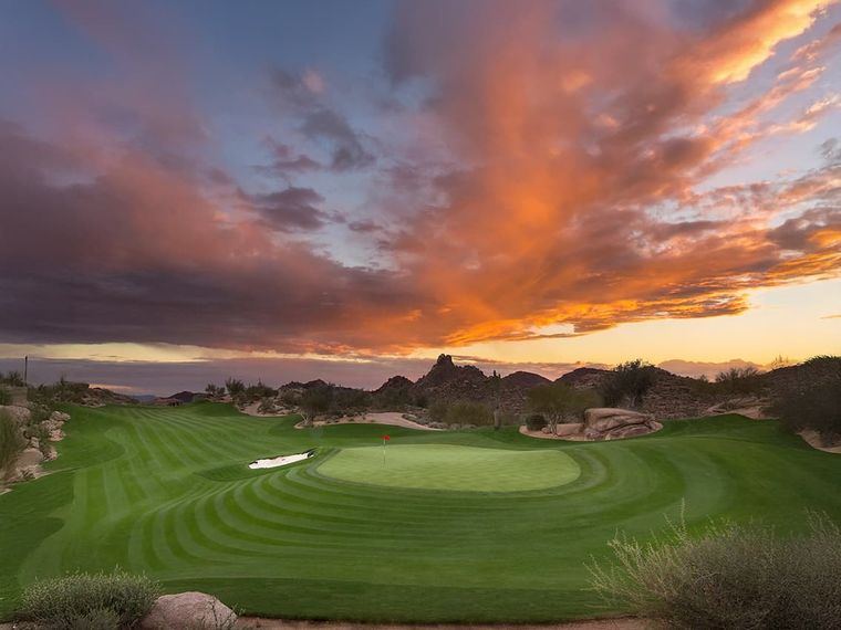 Golf green and flagstick under a vivid orange sunset over granite desert boulders
