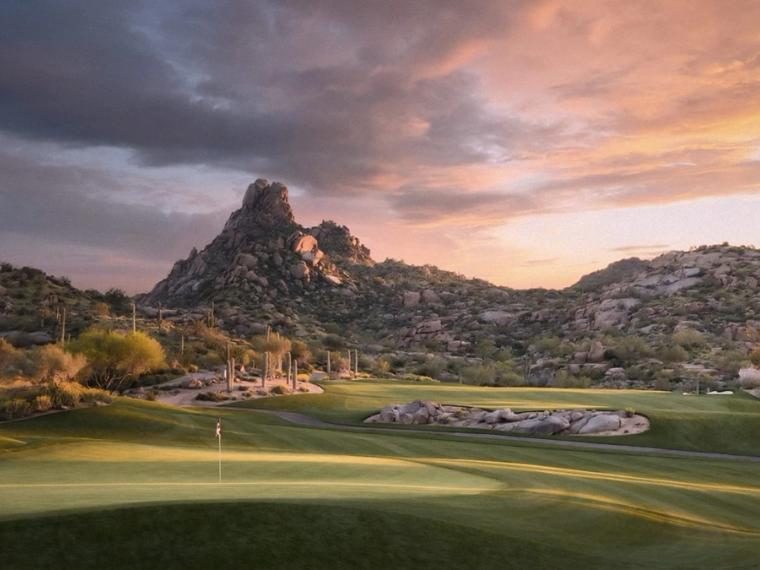 Putting green and flagstick in the foreground with a pointed rock peak and boulder-strewn hills behind, under an orange and violet sunset