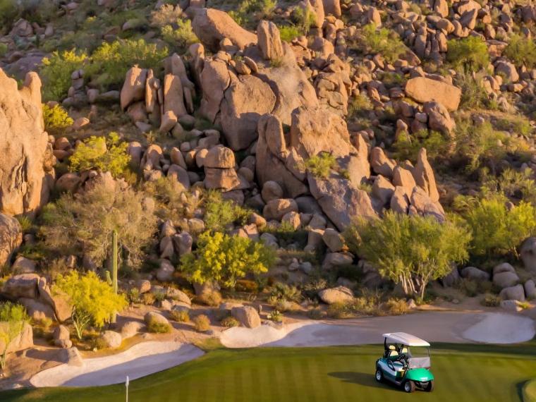 Golf cart parked beside a putting green and bunker at the foot of a steep boulder-covered hillside dotted with desert shrubs
