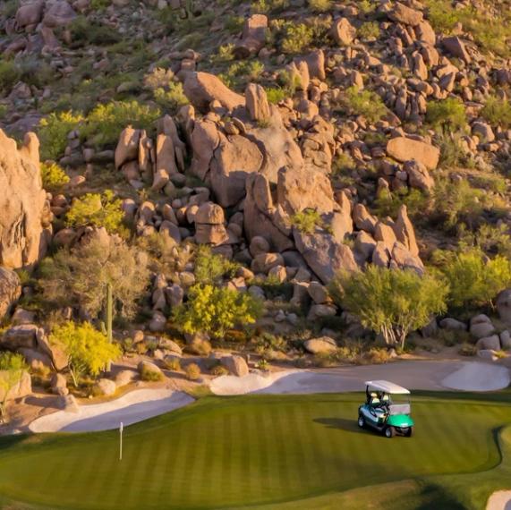 Golf cart parked beside a putting green and bunker at the foot of a steep boulder-covered hillside dotted with desert shrubs