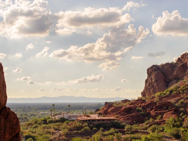 Red sandstone outcrops framing a view over desert homes to the valley floor and far mountains under scattered cloud