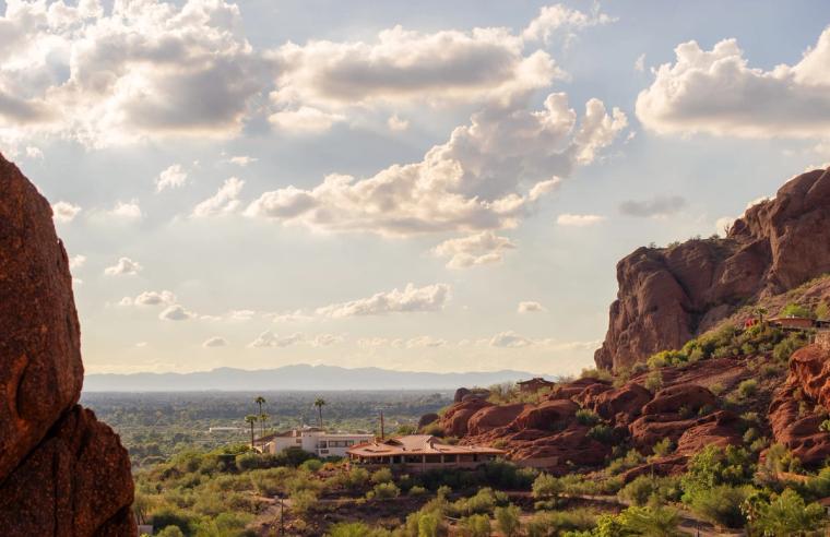 Red sandstone outcrops framing a view over desert homes to the valley floor and far mountains under scattered cloud