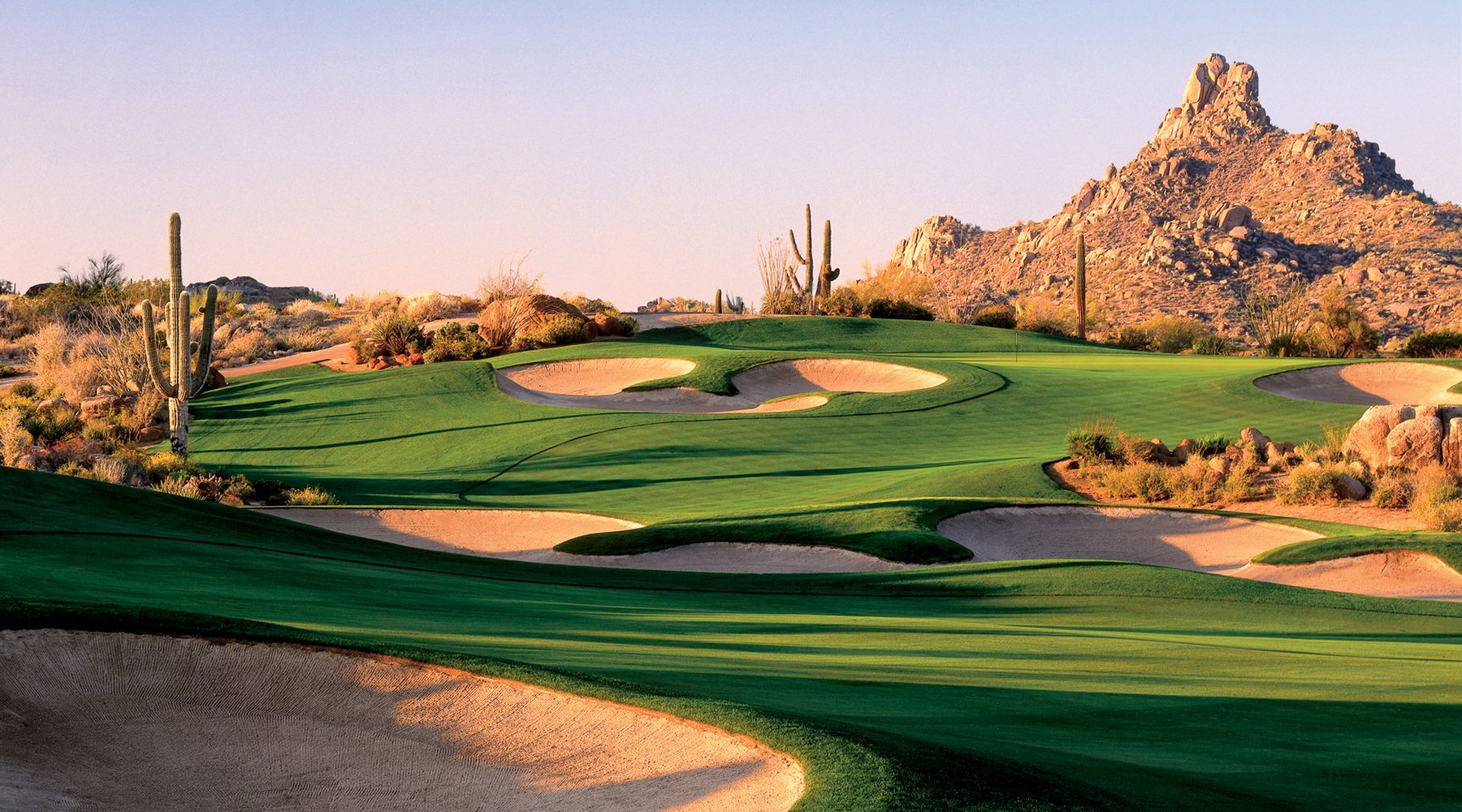 Golf green and sculpted bunkers ringed by saguaros below a granite boulder peak