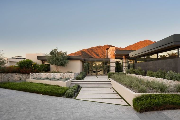 Flat-roofed stone and glass residence with paved entry walk below a sunlit ridge