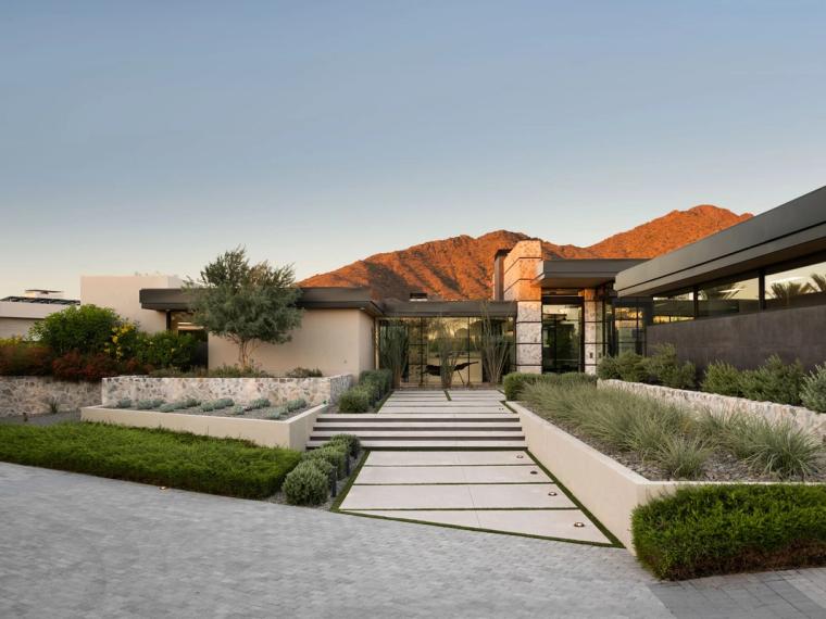 Flat-roofed stone and glass residence with paved entry walk below a sunlit ridge