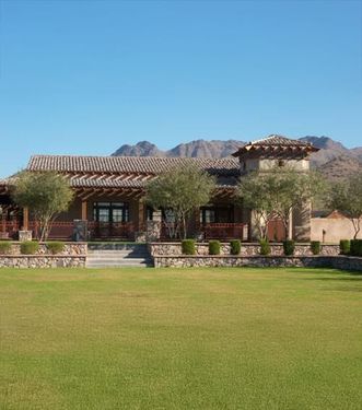 Low tile-roofed clubhouse with a stone base and deep shaded porch across a wide lawn, mountains rising behind