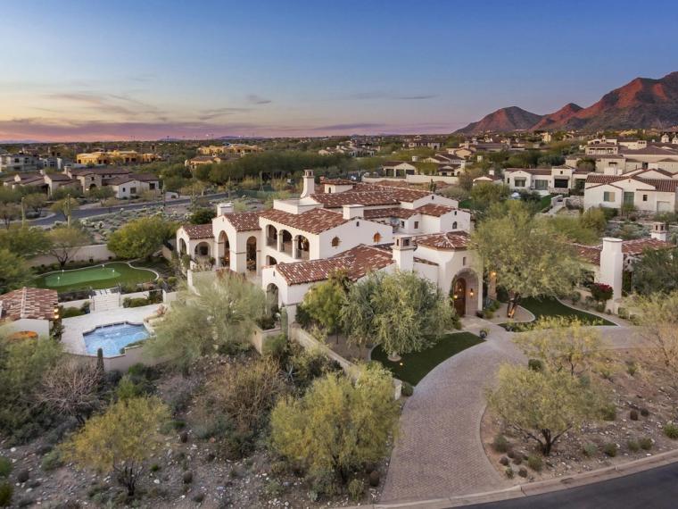 Aerial view at dusk of a tile-roofed house with a pool and lawn among neighboring homes, mountains catching last light