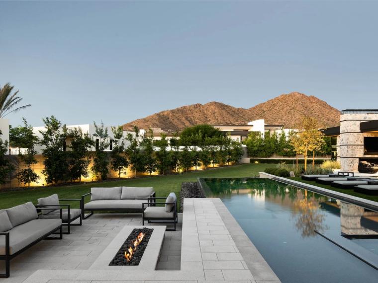 Contemporary home with lap pool and linear fire feature below a desert mountain at dusk