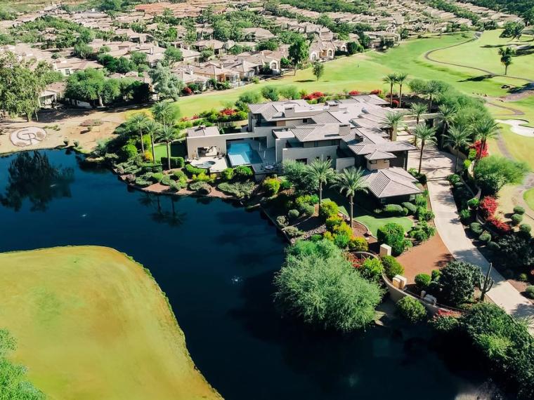 Aerial view of homes and a clubhouse around a dark lake, golf fairways and mature trees filling the frame