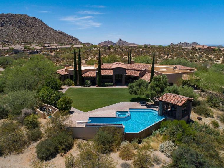 Aerial view of a desert estate with a pool, lawn and cypress trees, low mountains and a pointed peak in the distance