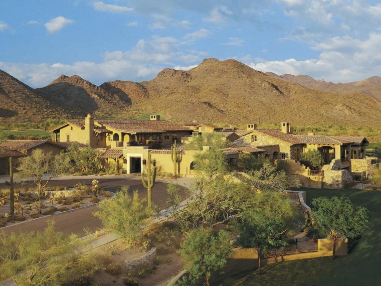 Low tile-roofed clubhouse in desert stone and stucco at the foot of a mountain, a curving drive and desert planting in front