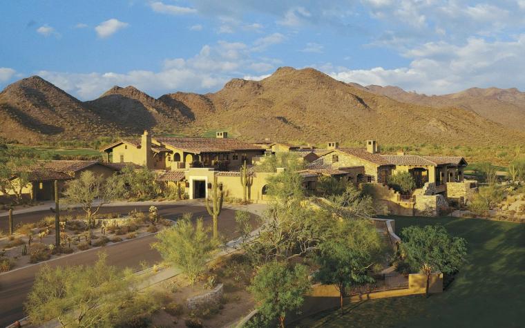 Low tile-roofed clubhouse in desert stone and stucco at the foot of a mountain, a curving drive and desert planting in front