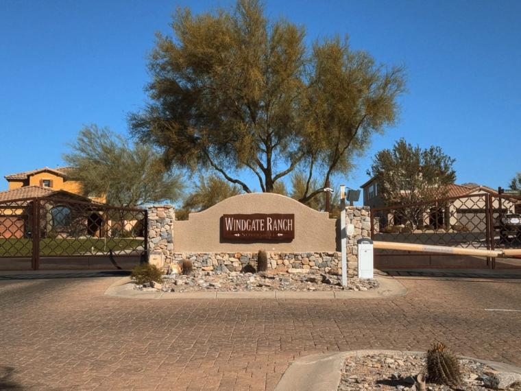 Stone entry monument lettered WINDGATE RANCH at a gated drive, a broad desert tree and tile-roofed buildings behind