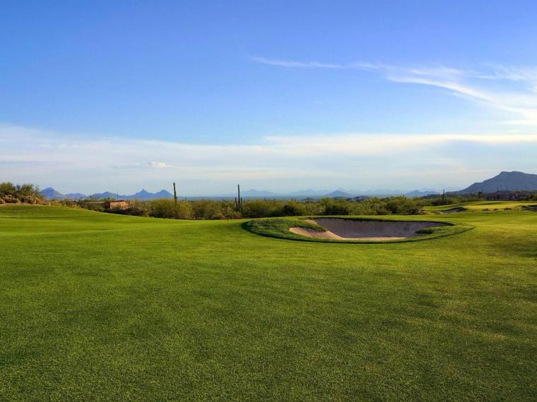 Wide green fairway and bunker running toward open desert, a dark peak on the far horizon under a pale blue sky