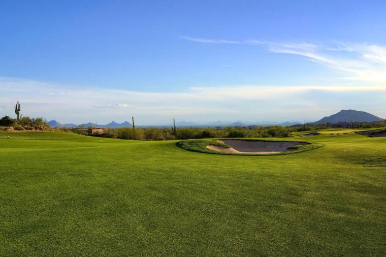 Wide green fairway and bunker running toward open desert, a dark peak on the far horizon under a pale blue sky