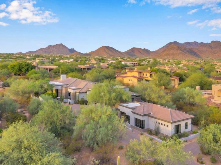 Aerial view of tile-roofed homes set among mature desert trees, a jagged mountain range across the horizon under a bright blue sky