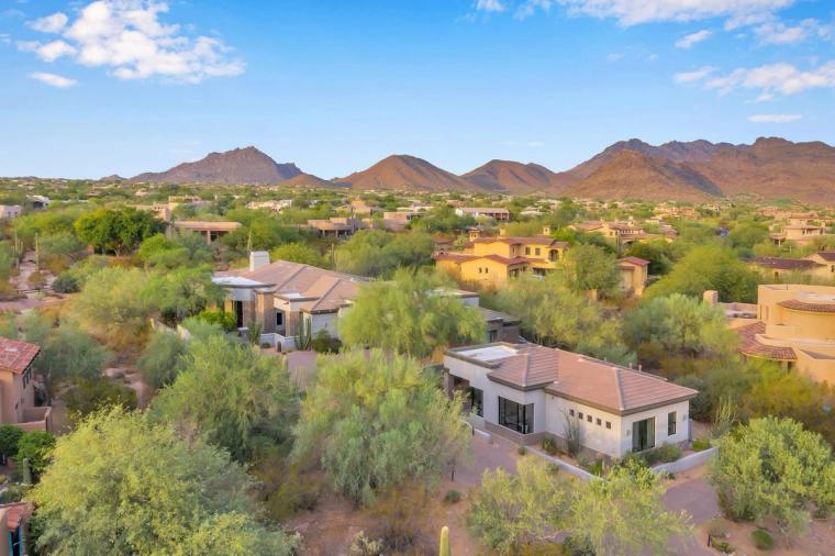 Aerial view of tile-roofed homes set among mature desert trees, a jagged mountain range across the horizon under a bright blue sky