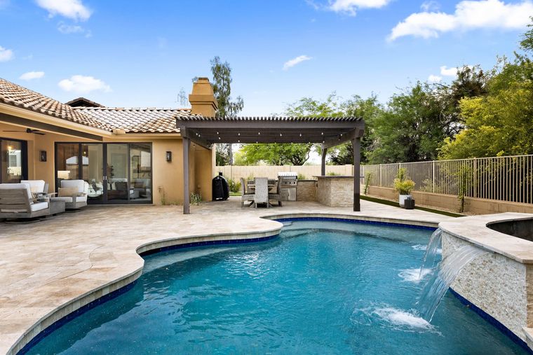 Backyard pool with waterfall spout, pergola and built-in grill beside a tile-roof home