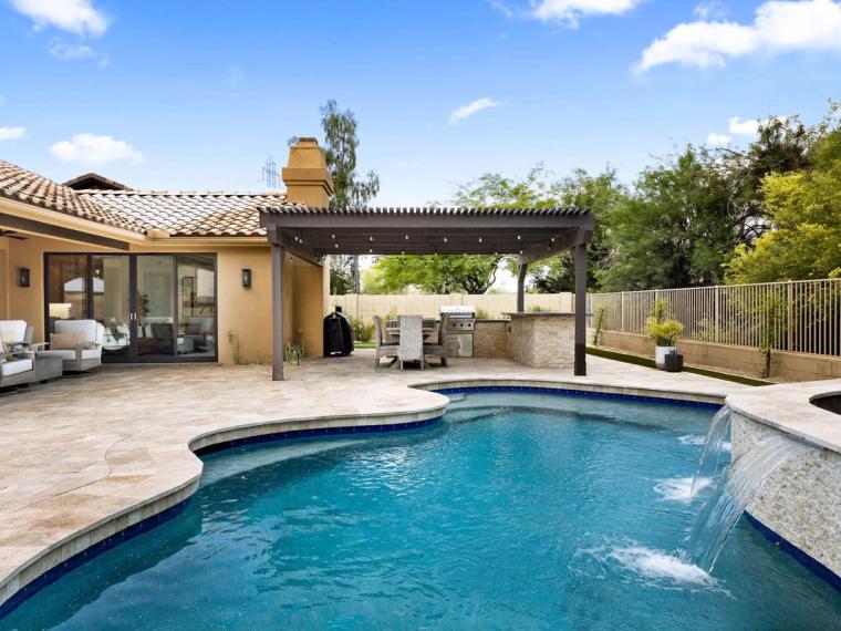 Backyard pool with waterfall spout, pergola and built-in grill beside a tile-roof home