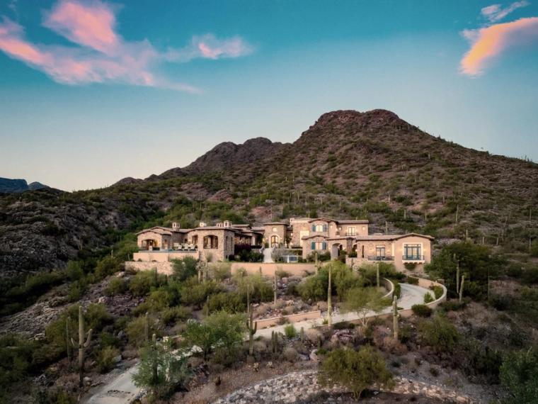 Large stucco house on a desert slope below a rounded peak, saguaro and scrub across the hillside under a pink-streaked sky