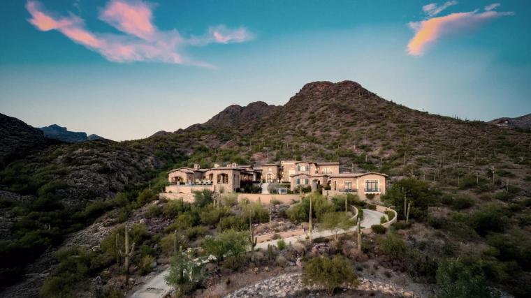 Large stucco house on a desert slope below a rounded peak, saguaro and scrub across the hillside under a pink-streaked sky