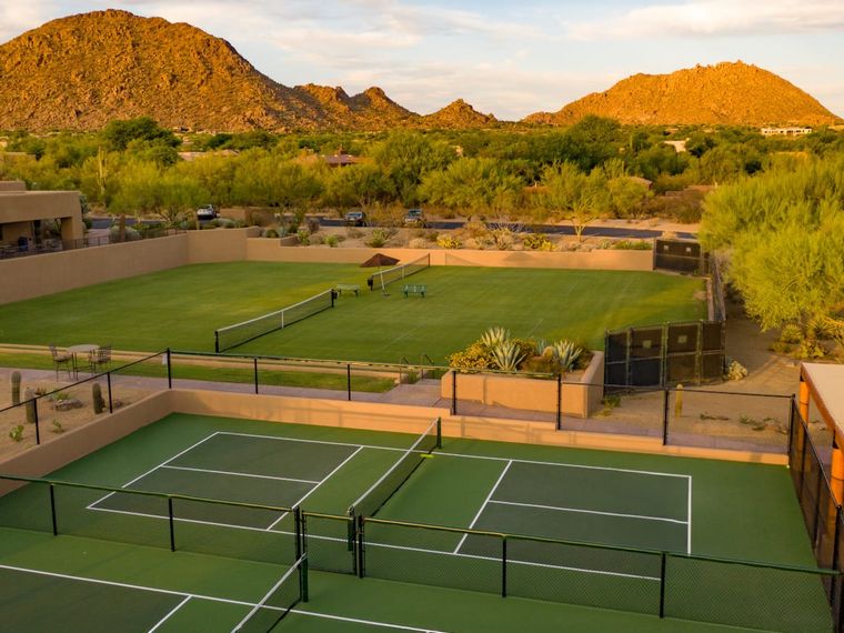Elevated view of tennis and pickleball courts terraced into the desert below sunlit hills and a cloud-streaked sky