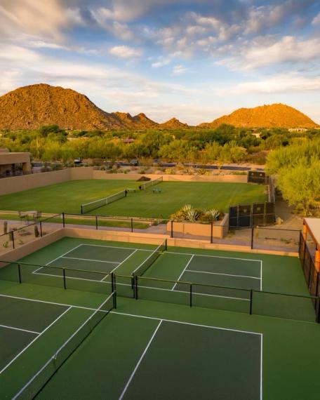 Elevated view of tennis and pickleball courts terraced into the desert below sunlit hills and a cloud-streaked sky