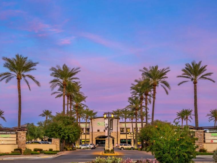 Low commercial building set back from a street behind tall palms and flowering planting, under a pink and blue dusk sky