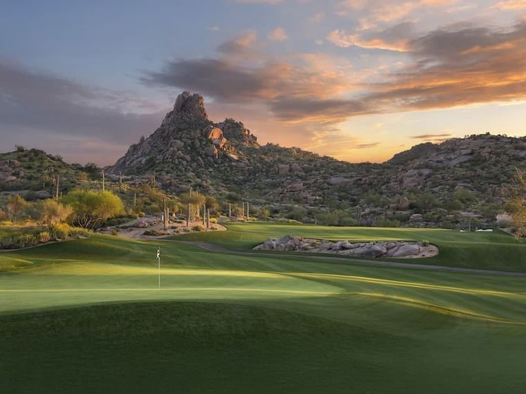 Fairway and green at dusk below a jagged rock summit, boulders and desert brush catching the last light