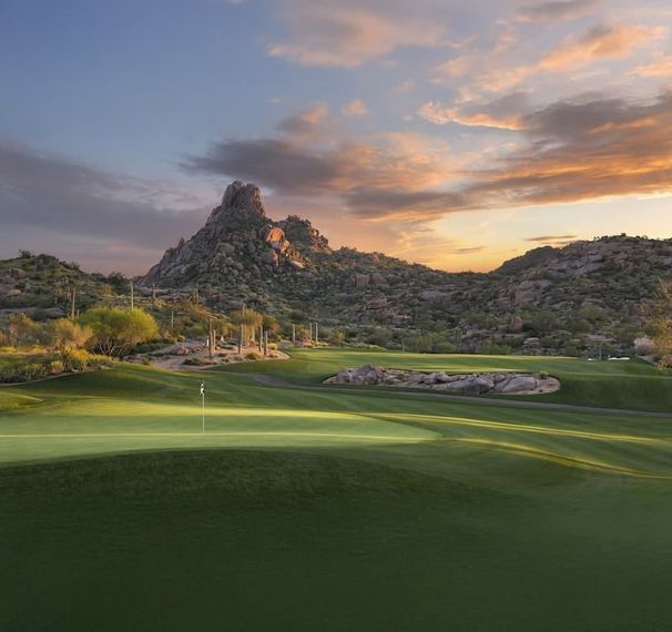 Fairway and green at dusk below a jagged rock summit, boulders and desert brush catching the last light