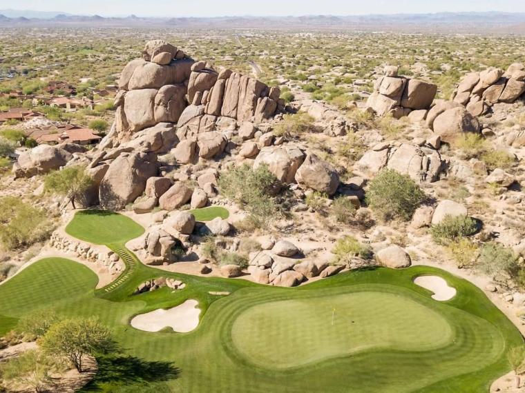 Aerial view of a green and bunkers wrapped around a large outcrop of granite boulders, open desert stretching beyond