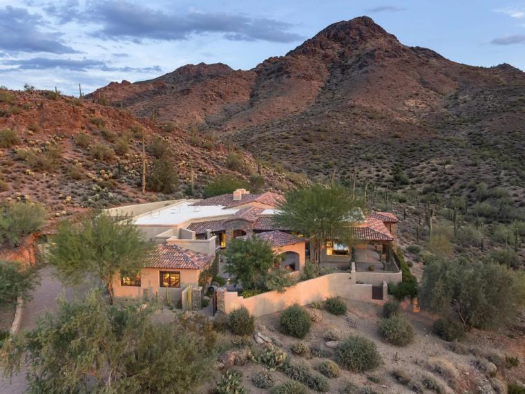 Tile-roofed house tucked into a desert hillside below a steep rocky peak, saguaro and brush across the slope