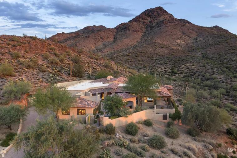 Tile-roofed house tucked into a desert hillside below a steep rocky peak, saguaro and brush across the slope