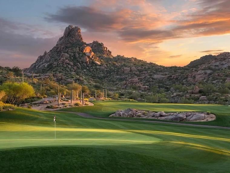 Putting green and flagstick in the foreground with a pointed rock peak and boulder-strewn hills behind, under an orange and violet sunset
