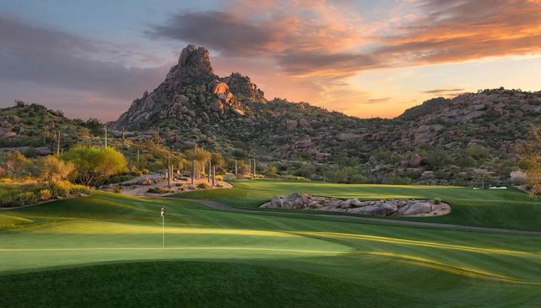 Putting green and flagstick in the foreground with a pointed rock peak and boulder-strewn hills behind, under an orange and violet sunset