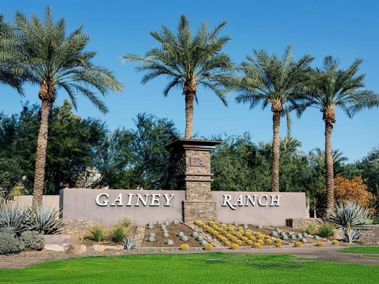 Stone and stucco entry wall lettered GAINEY RANCH, flanked by tall date palms and beds of agave and flowering shrubs