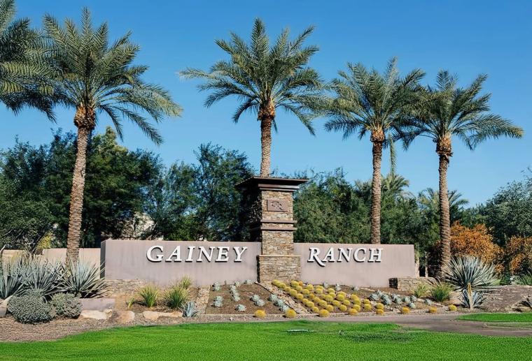 Stone and stucco entry wall lettered GAINEY RANCH, flanked by tall date palms and beds of agave and flowering shrubs