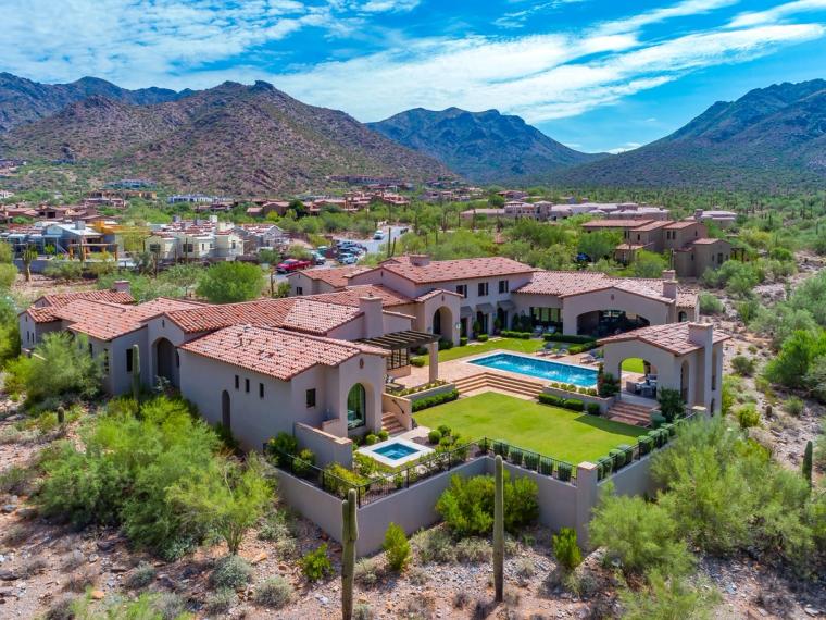 Aerial view of a tile-roofed stucco house with a pool and walled lawn, set in desert planting below a mountain range