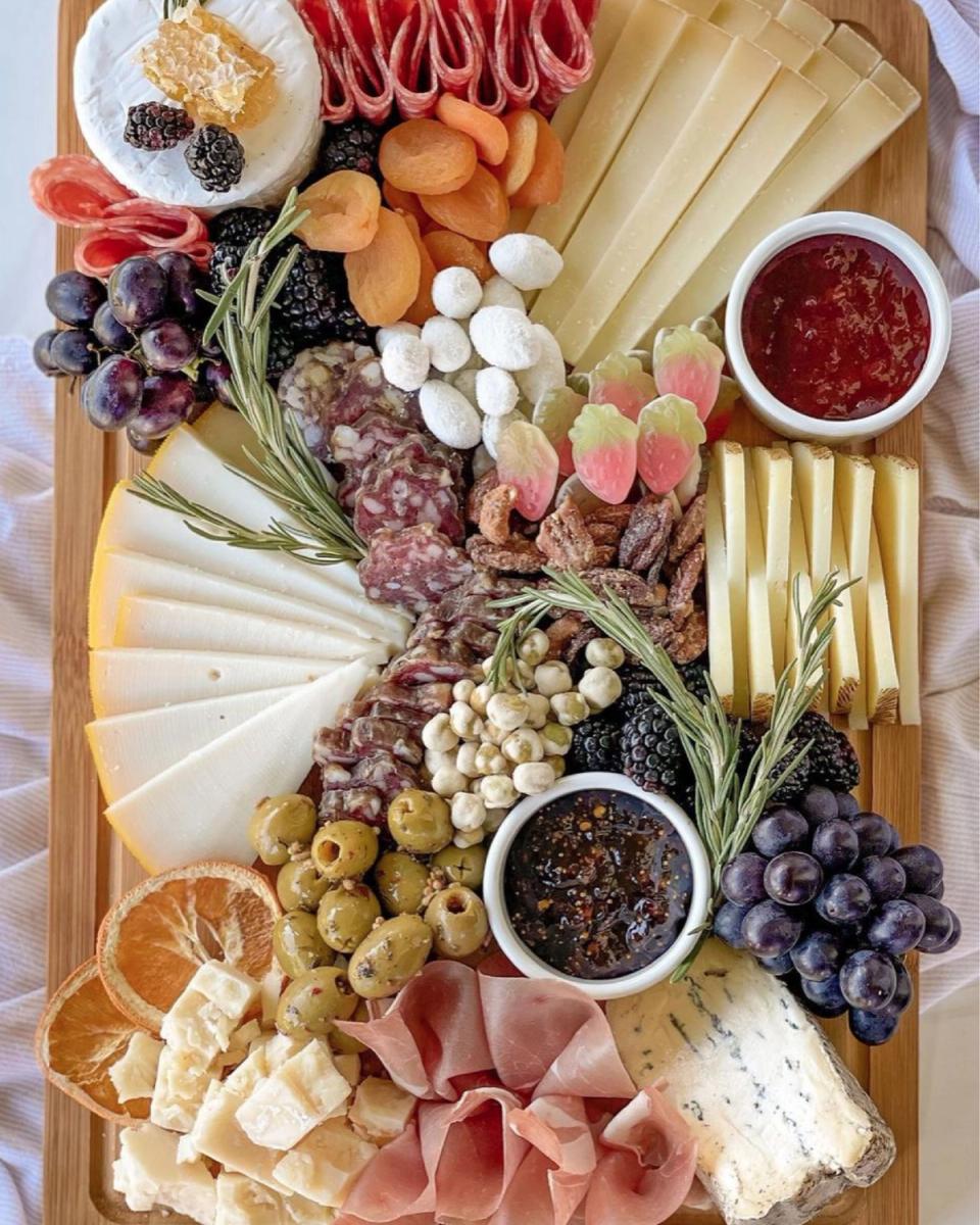 Overhead view of a wooden charcuterie board fanned with cured meats, sliced cheeses, olives, dried fruit, and rosemary sprigs