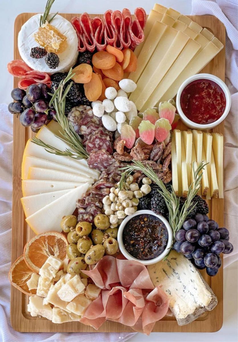 Overhead view of a wooden charcuterie board fanned with cured meats, sliced cheeses, olives, dried fruit, and rosemary sprigs